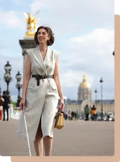 Muriel Joubert, coach entrepreneur, souriante marchant en extérieur sur un pont parisien, tenant une canne blanche et un sac, avec des monuments en arrière-plan.